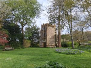 Cerne Abbey, the abbots porch in a lush garden, framed by trees, with benches and a grassy lawn in the foreground.