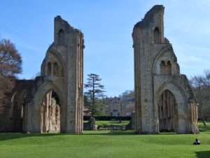 Ruined Gothic arches of an Glastonbury abbey with a green lawn and a few visitors in the distance.
