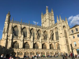 Front view of Bath Abbey, a Gothic stone church with tall lancet windows and decorative pinnacles under a clear blue sky; people seated along the plaza in front.