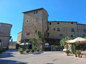 Stone medieval building with a tall tower in a sunny town square, outdoor cafe seating with umbrellas, and potted plants.