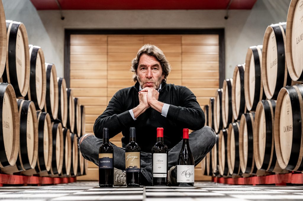 Man sitting cross‑legged between wine barrels in a cellar, arms resting on his knees, with five wine bottles lined up in front of him.