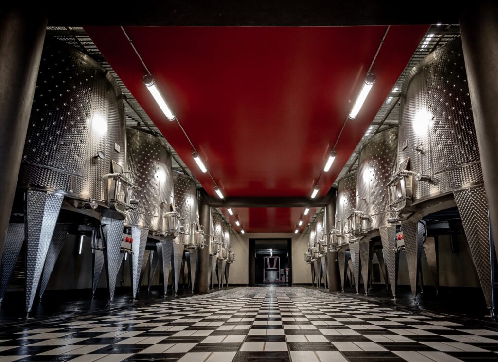 La Massa winery or brewery cellar with stainless steel fermentation tanks along a hallway and a black-and-white checkered floor under a red ceiling.