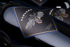 Close-up of a dark wine bottle with a black label reading 'VARVARA' and a crest, framed by a blue-gold border.